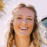 A woman with blonde hair smiling at the camera with a sunny background. 